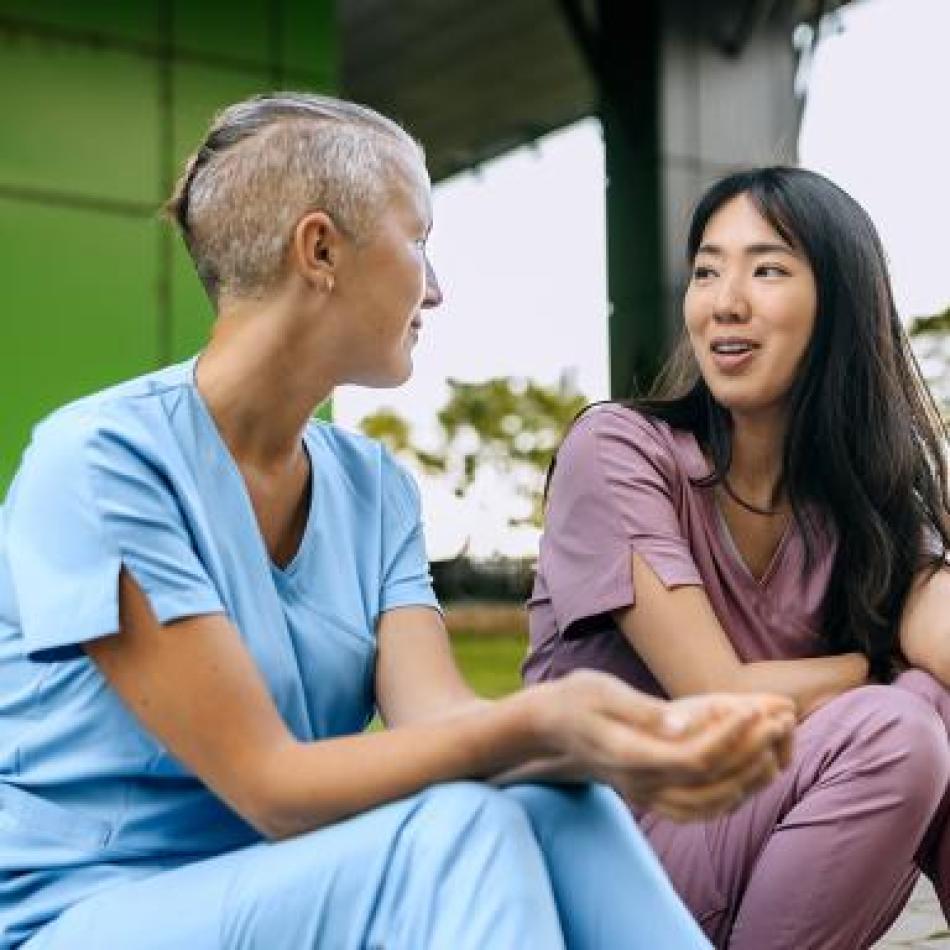 Two nurses in scrubs chat outdoors. One with short hair, one gesturing whilst speaking.
