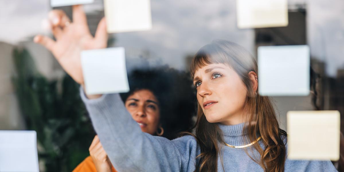 Photograph: Two women intently arranging yellow sticky notes on a glass screen.